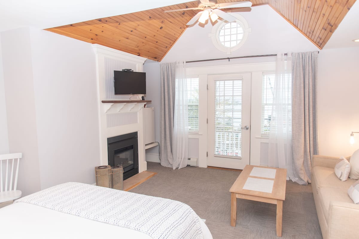 A bedroom with a bed, a wooden ceiling, a television, a fireplace, and a door with curtains.