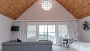 A room with a sloped wooden ceiling, a round skylight, a television mounted on the wall, a sofa, a bed, a dining table with chairs, and a window with blinds. - Inn On Peaks Island (Peaks Island)