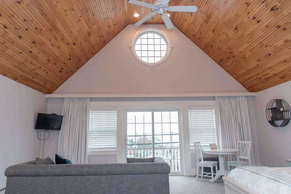 A room with a sloped wooden ceiling, a round skylight, a television mounted on the wall, a sofa, a bed, a dining table with chairs, and a window with blinds.