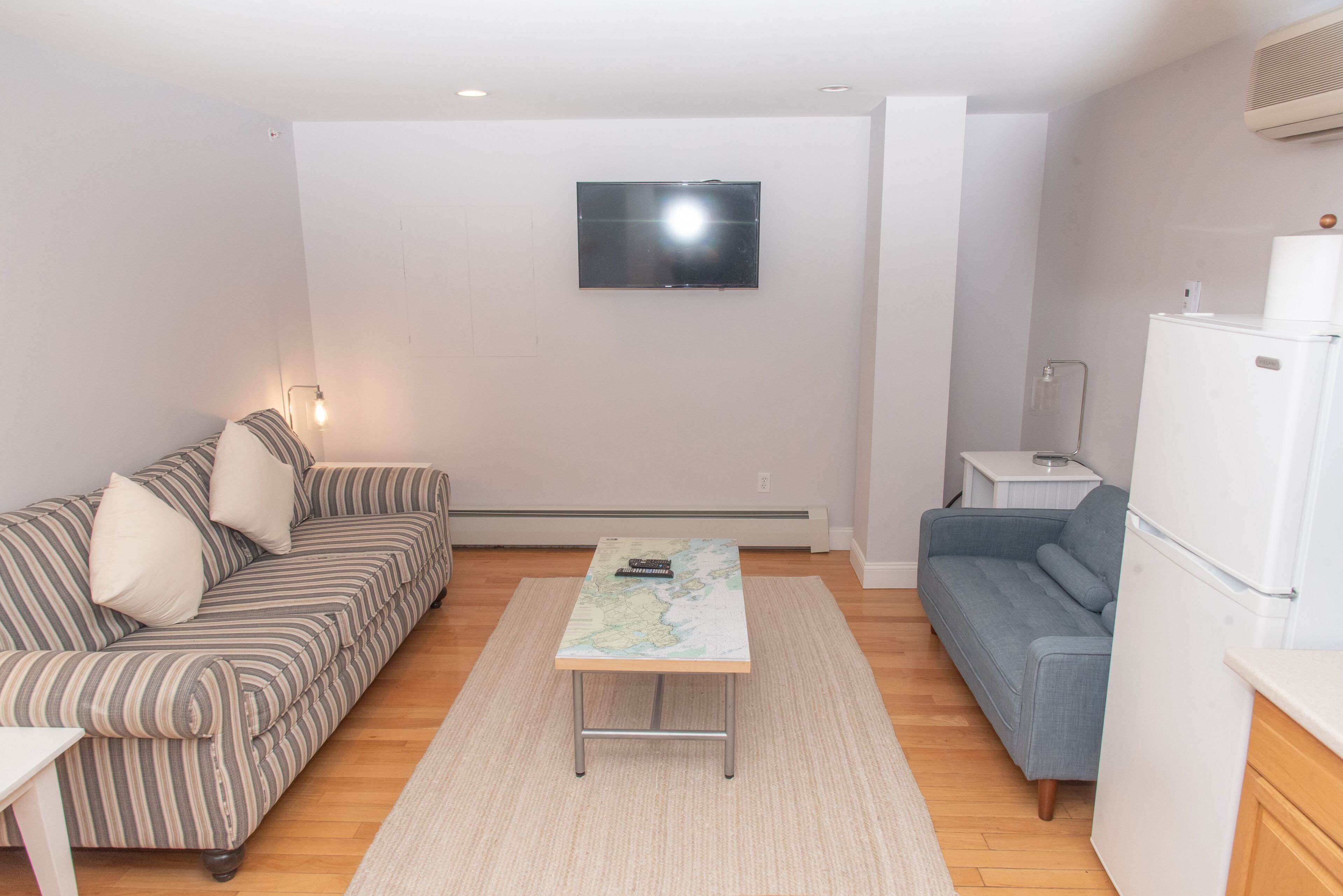 Ein kleiner, moderner Wohnraum mit einer Couch, einem Couchtisch mit einer Karte, einem an der Wand befestigten Fernseher und einem Kühlschrank.
