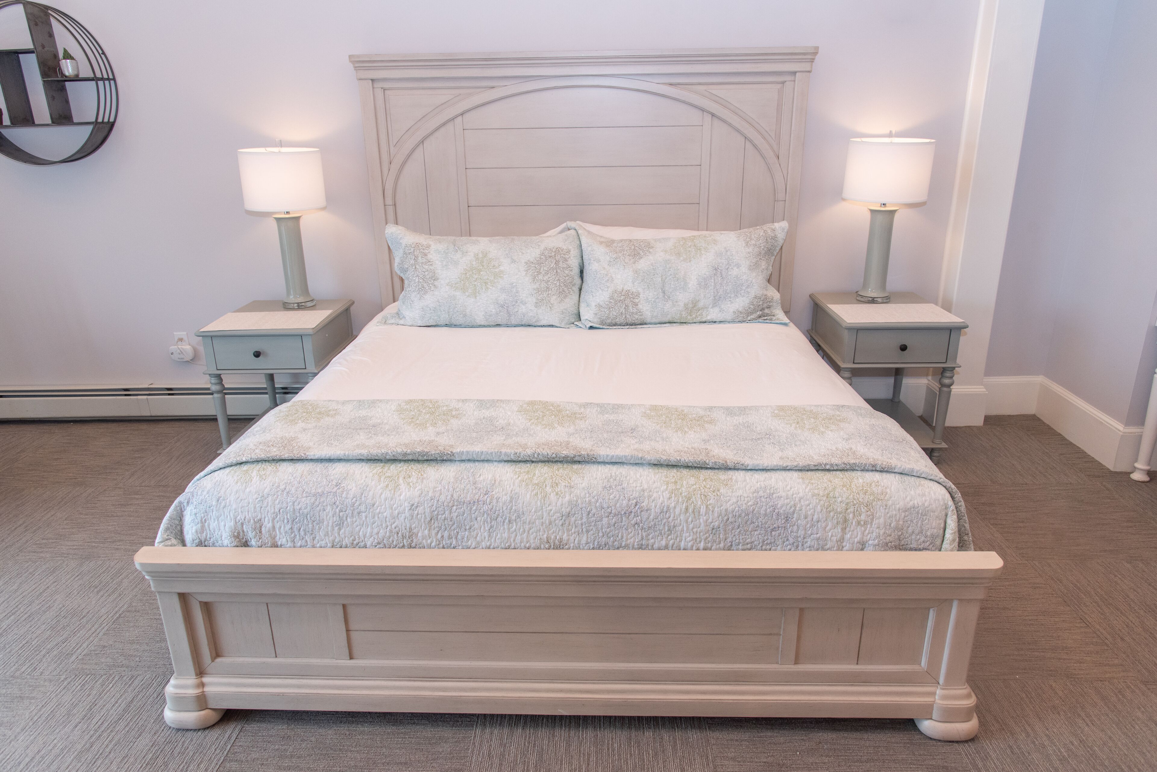 Cama bem arrumada com cabeceira, duas mesas de cabeceira com luminárias e prateleira fixada na parede.