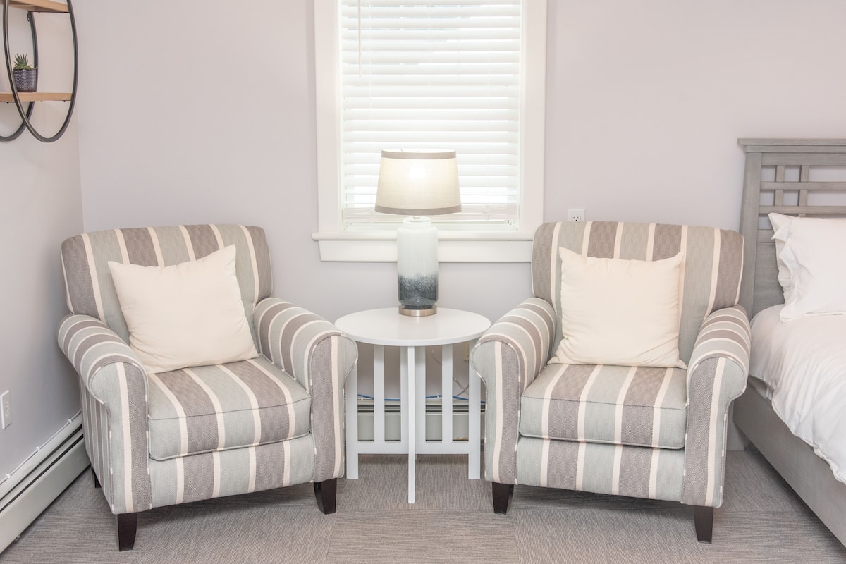 A room with two striped armchairs, a small round table with a lamp, and a bed with a headboard.