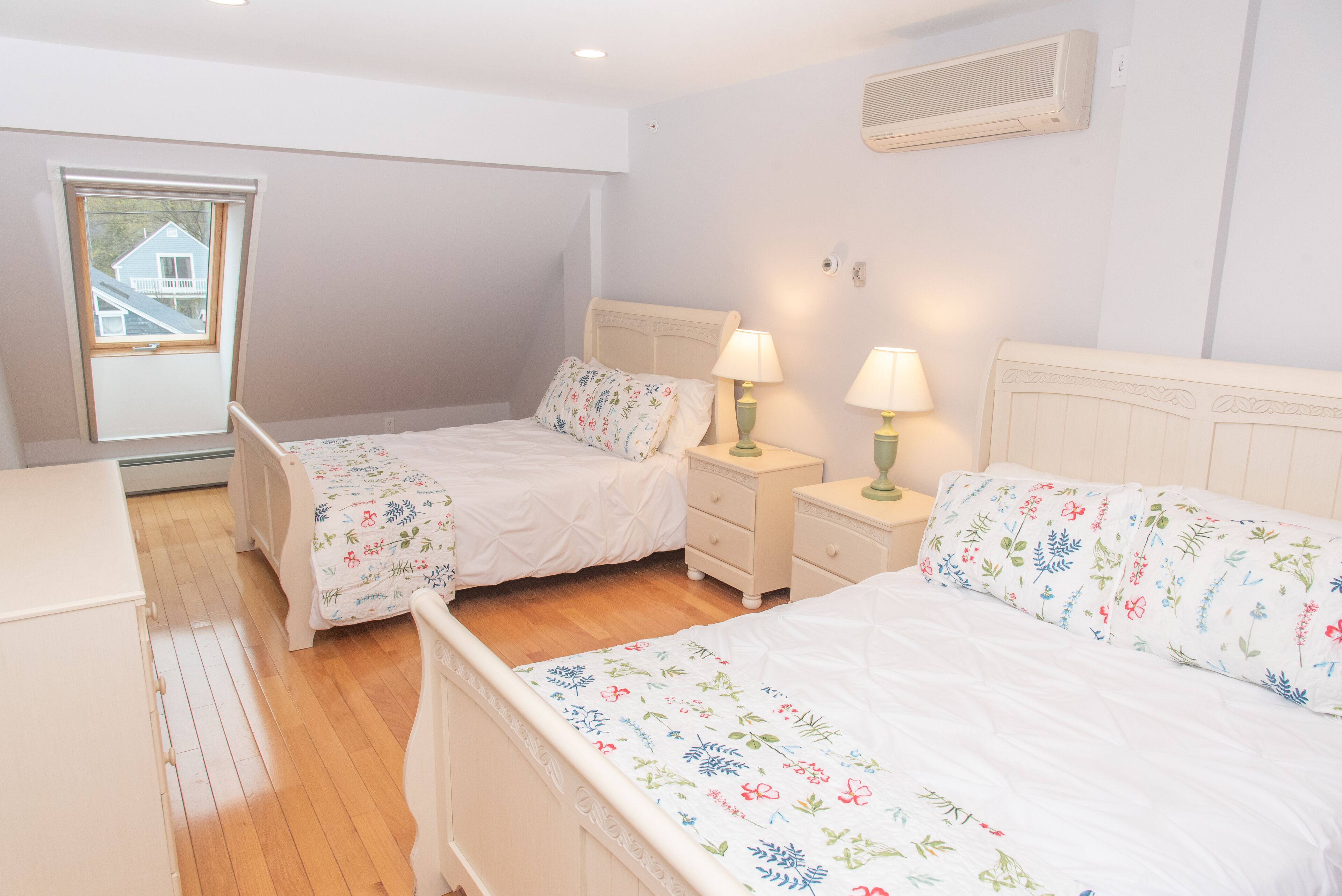 A bedroom with two single beds, wooden flooring, a window with a view, and a wall-mounted air conditioner.