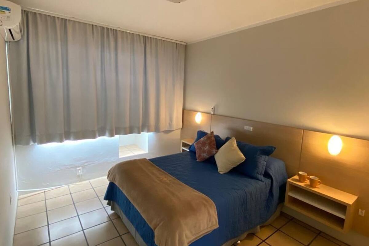 A bedroom with a bed, curtains, a bedside table, and a wall-mounted shelf.
