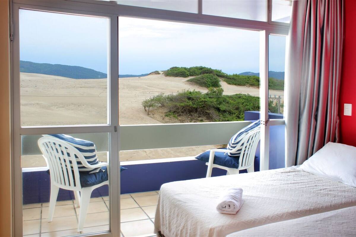 A hotel room with a bed, a window with a view of the beach, and two chairs with cushions.