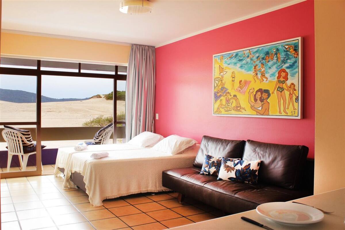 A hotel room with a bed, a brown leather sofa, a painting on the wall, and a view of the desert through large windows.