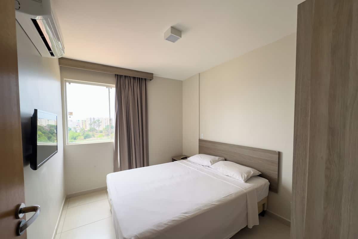 A hotel room with a bed, a TV mounted on the wall, a window with curtains, and an air conditioning unit.