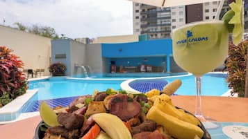 Ein Esstisch mit verschiedenen gegrillten Fleischsorten und Gemüse, einem Cocktailglas sowie einem Pool im Hintergrund.