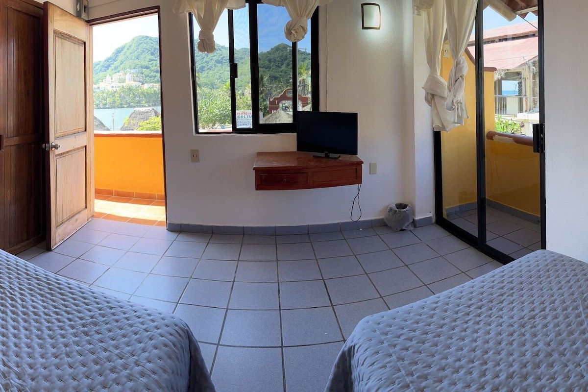 Pasillo de hotel con dos camas, un televisor y vista a un paisaje montañoso a través de una ventana.