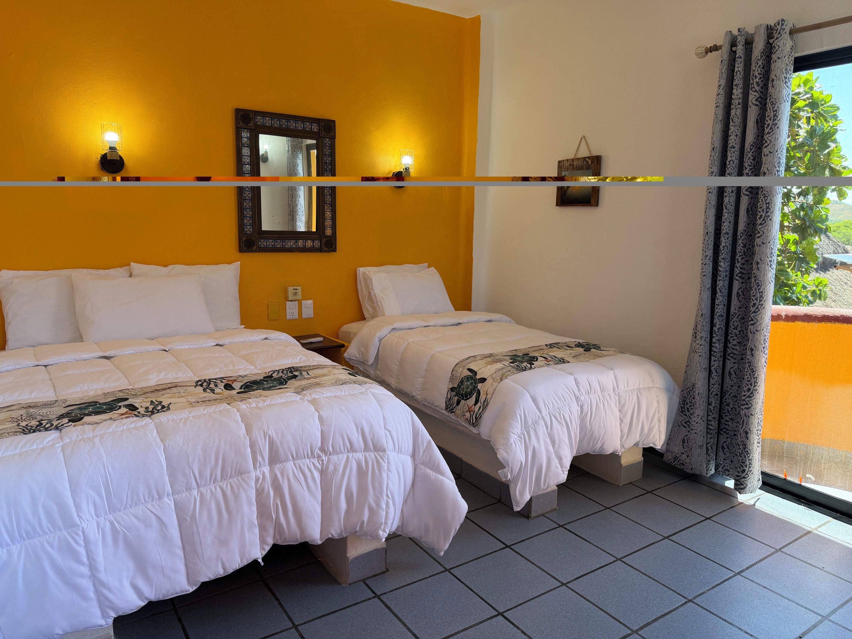 A hotel room with two beds, a large mirror, wall sconces, and a view of greenery outside.