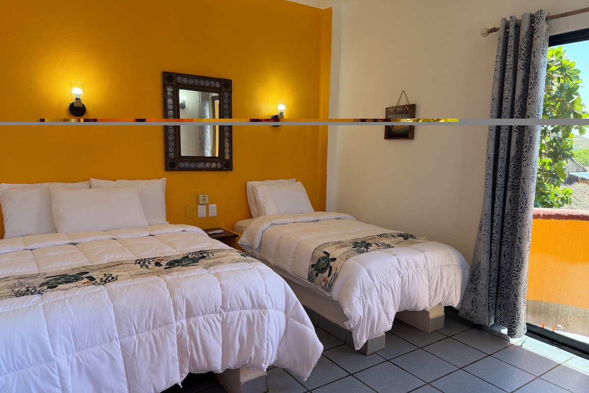 Habitación de hotel con dos camas, un espejo grande, apliques de pared y vista a zonas verdes.