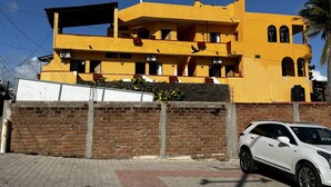 Un immeuble jaune à plusieurs étages, avec un mur de briques à l’avant, une voiture blanche garée à proximité et un ciel bleu dégagé.