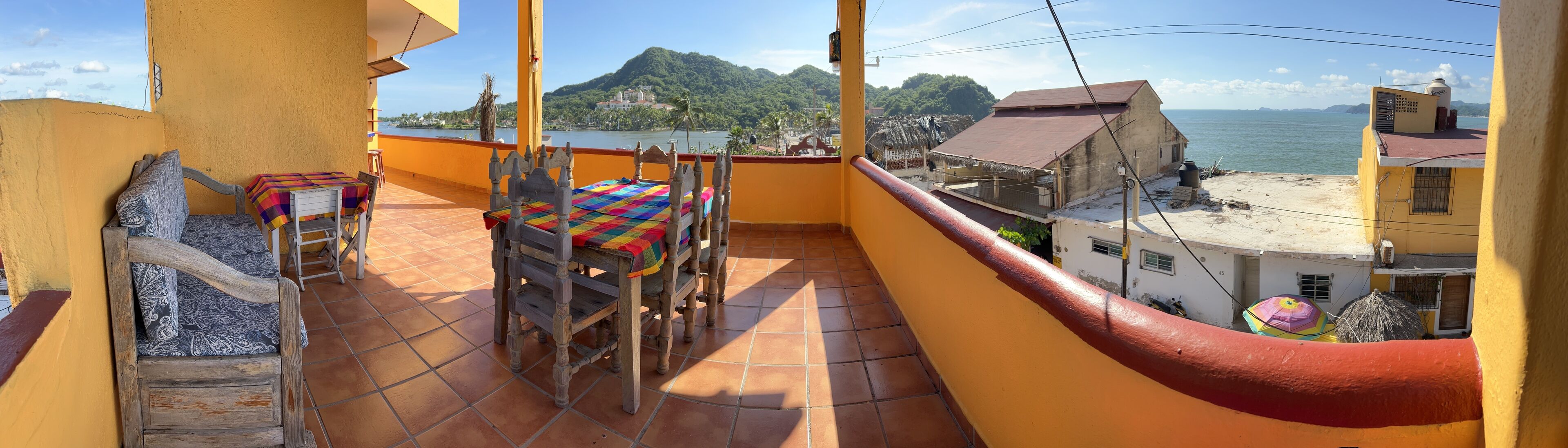 A balcony with a wooden bench and table, a tiled floor, and a view of a coastal area with buildings and greenery.