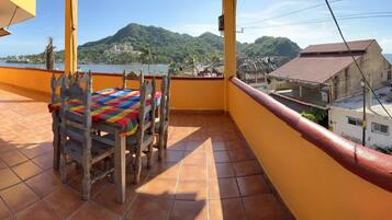 A balcony with a wooden bench and table, a tiled floor, and a view of a coastal area with buildings and greenery.