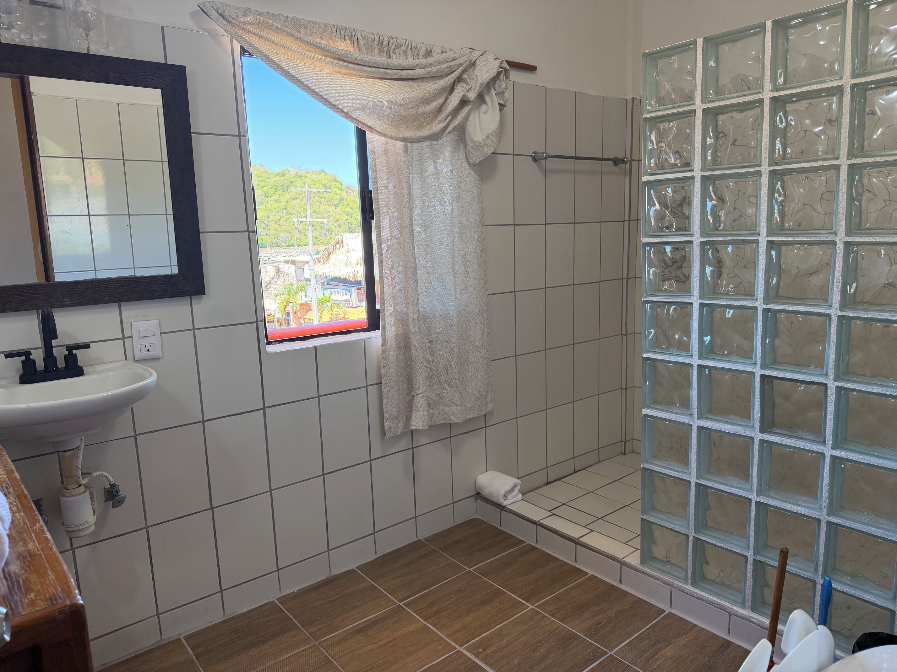 A bathroom with a white sink, a mirror, a window with a view of greenery, and a shower with frosted glass panels.