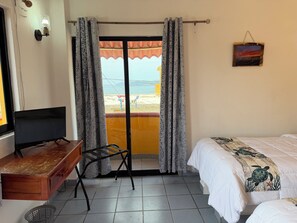 Una habitación con cama, escritorio con televisor, silla y vista a la playa a través de cortinas.