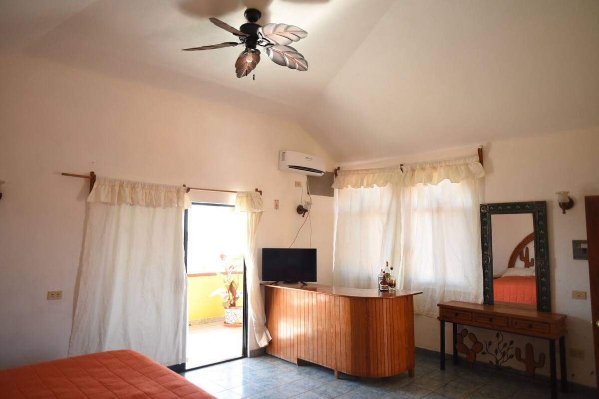 Una habitación con cama, escritorio, ventilador de techo y ventana con cortinas.
