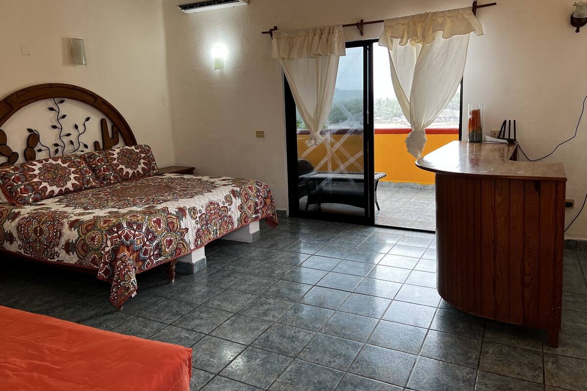 Un dormitorio con cama, un escritorio de madera, un balcón con cortinas y un equipo de aire acondicionado.