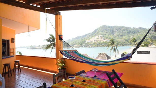 Un balcon avec un hamac, une table recouverte d’une nappe colorée et une vue sur une plage et des montagnes.