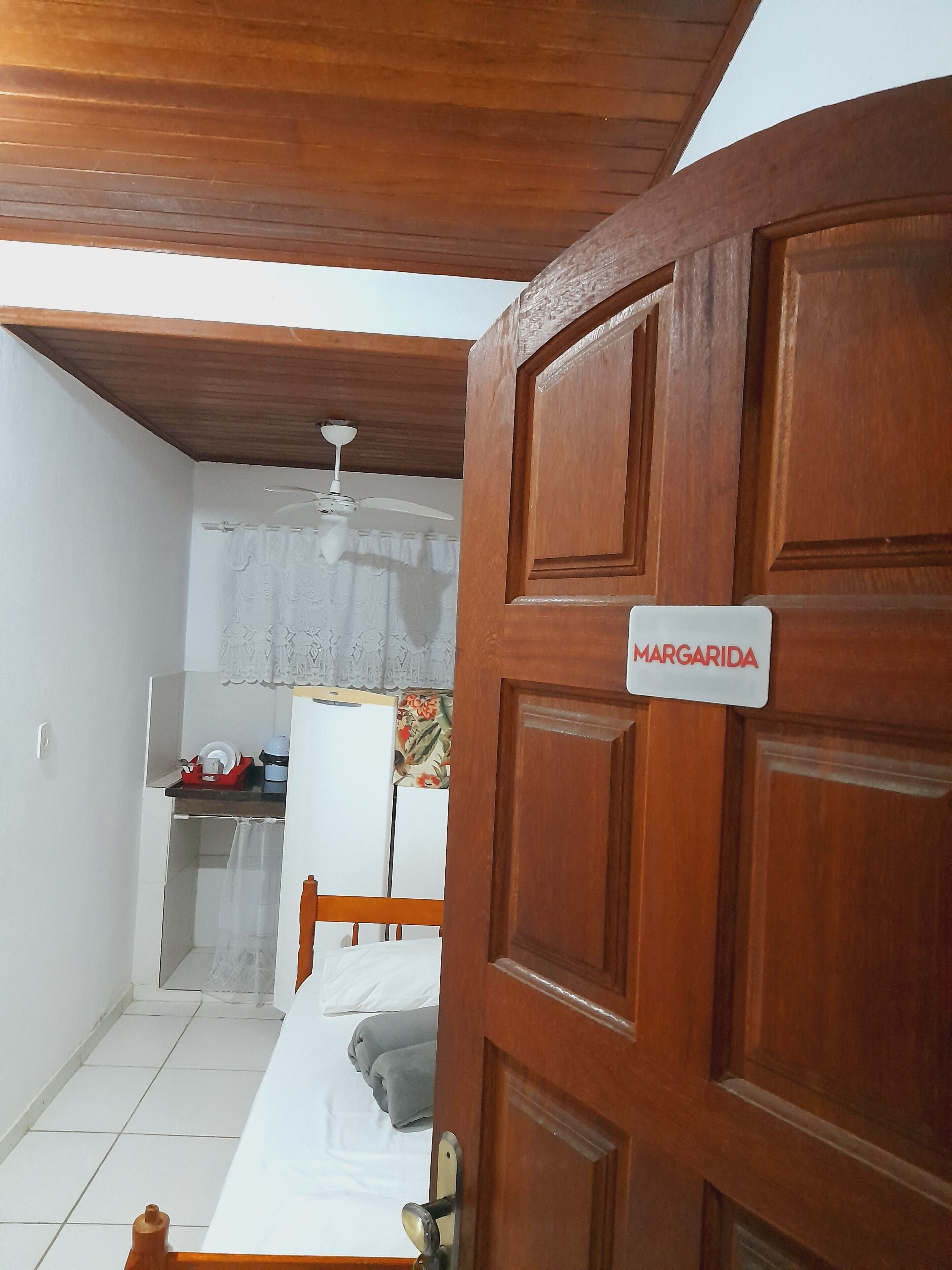 A room with a wooden door labeled "MARGARIDA," a bed, a small table with a white lamp, and a ceiling fan.