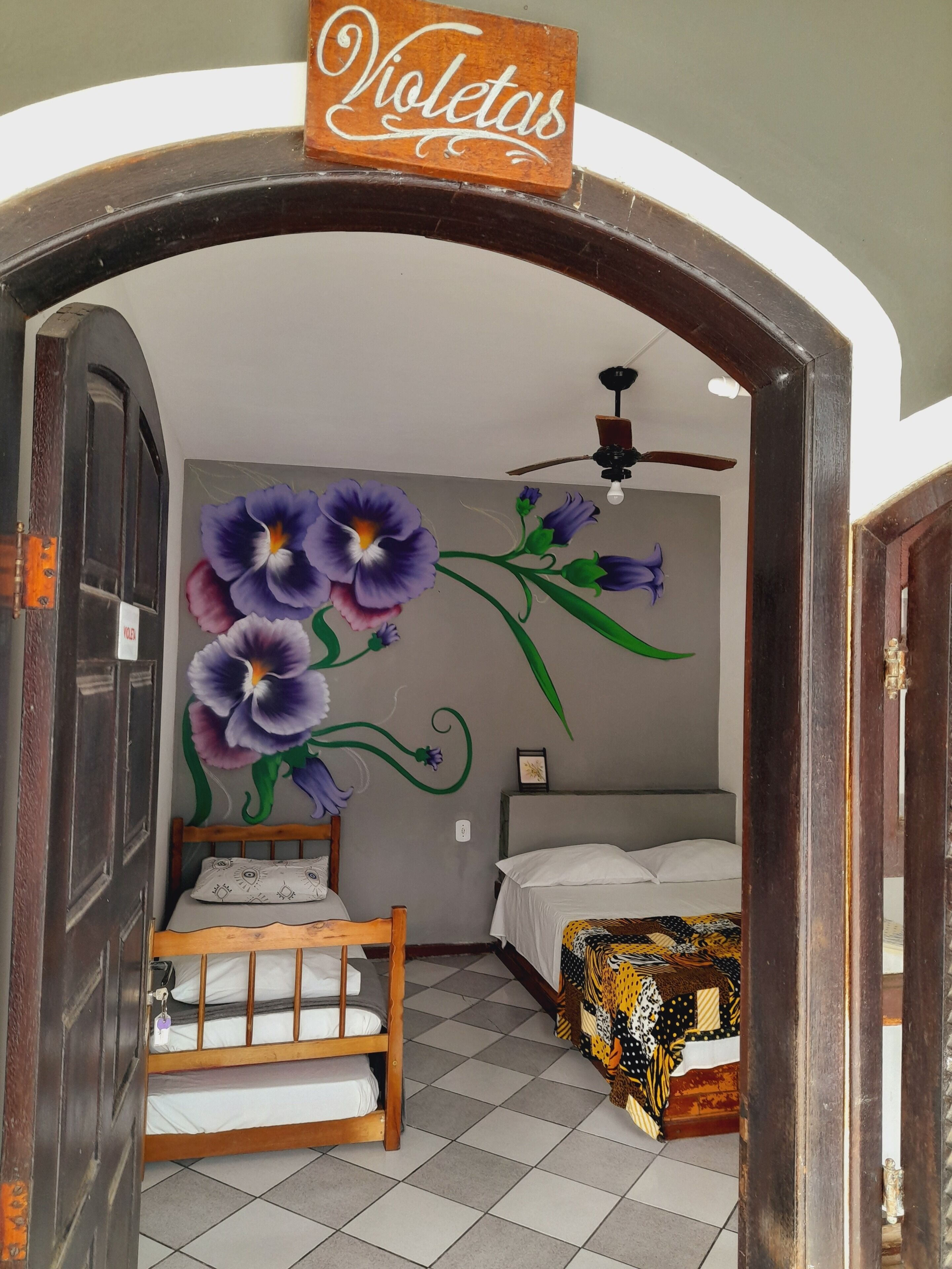 Ein Zimmer mit einem Bett, einem kleinen Tisch, einem Deckenventilator und einer Wand mit floralem Wandgemälde.
