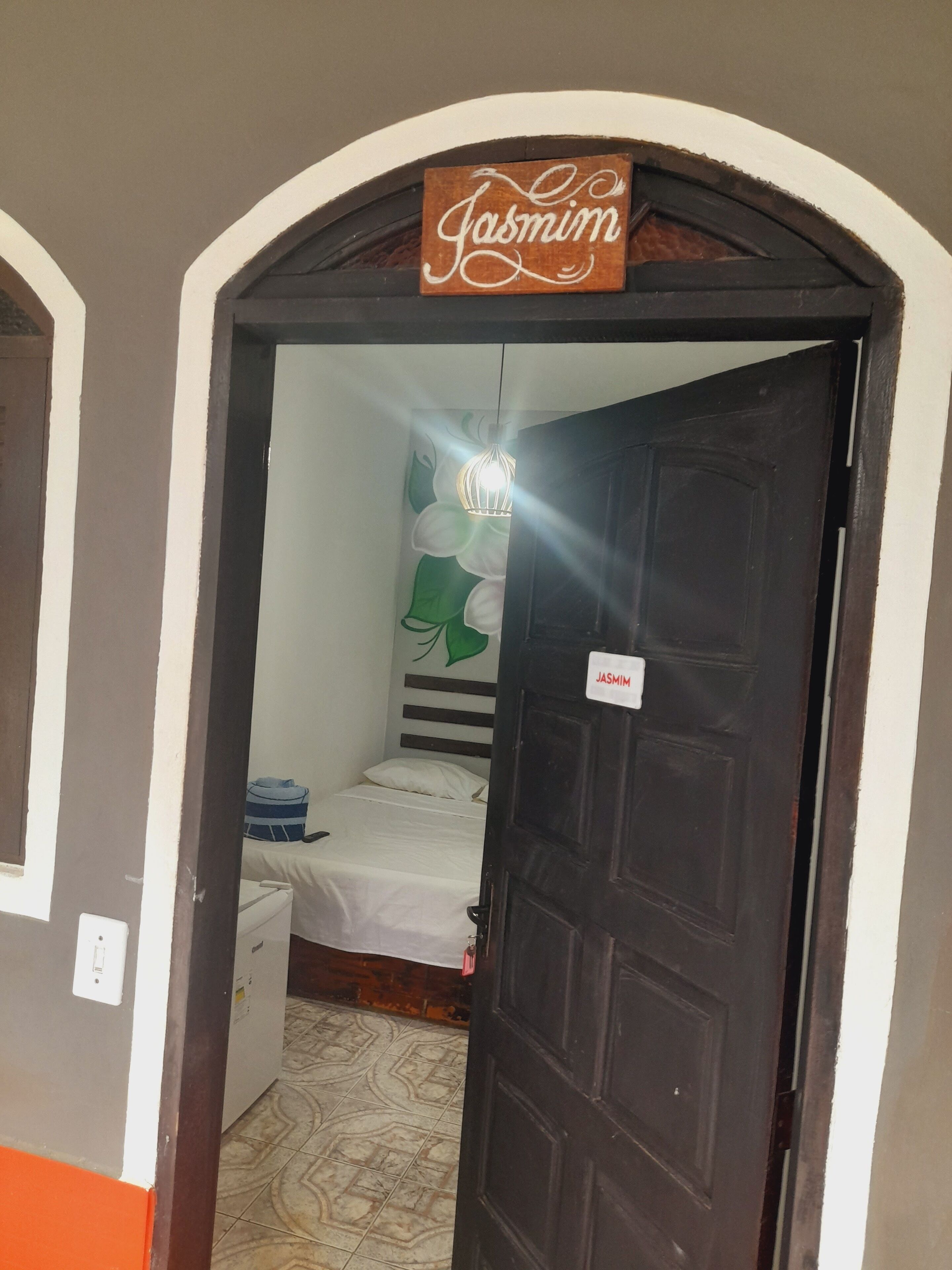 Una habitación con una cama, una lámpara fijada a la pared y una puerta con un letrero que dice 'Jasmin'.