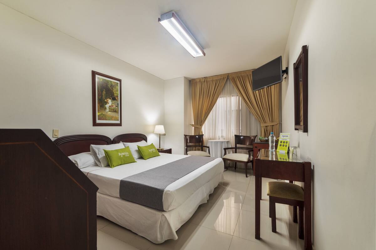 A hotel room with a bed, bedside table, lamp, chair, desk, and a television on the wall.