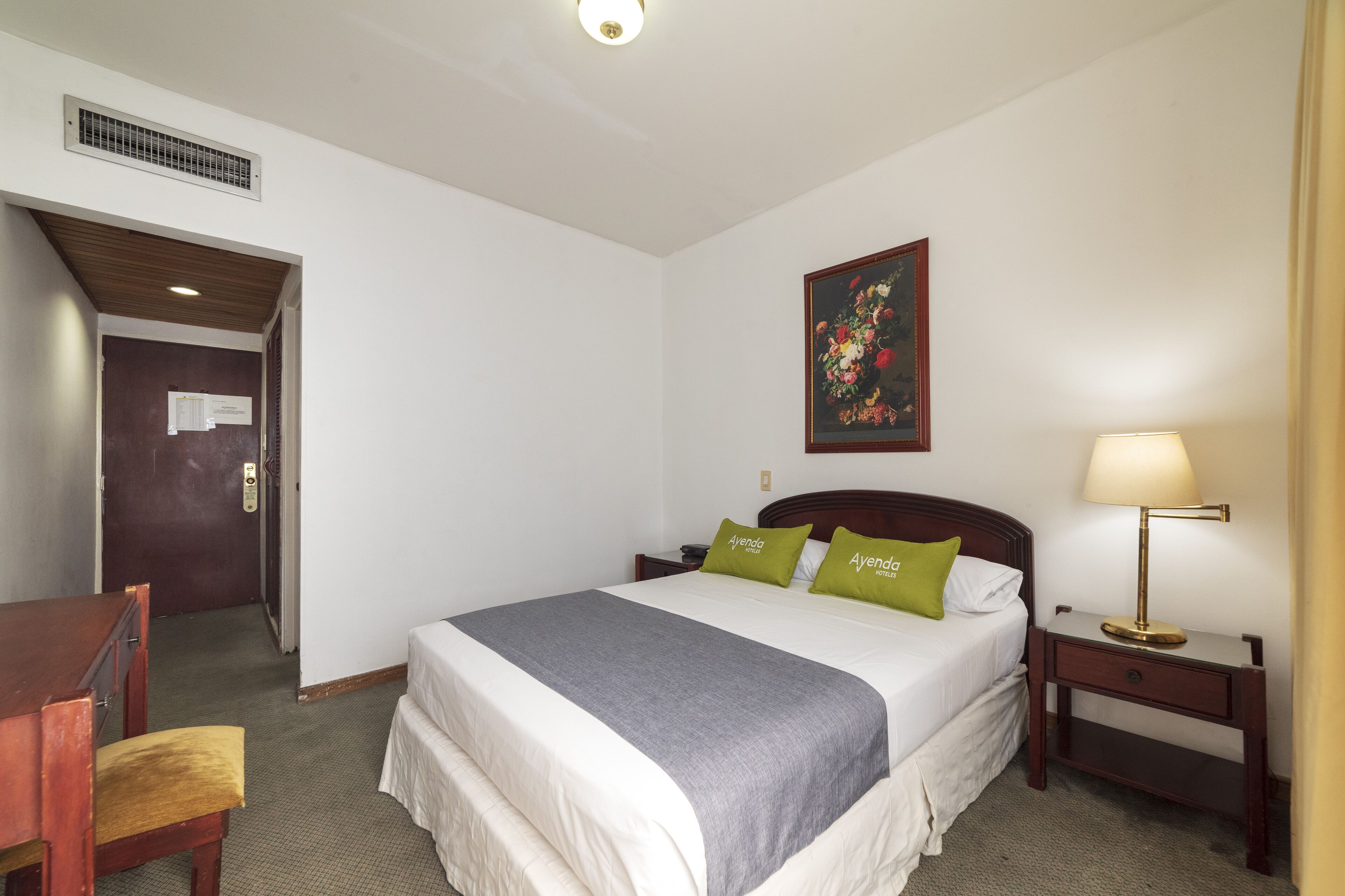 A hotel room with a bed, a desk, a chair, a nightstand with a lamp, a painting on the wall, and a door leading to another room.