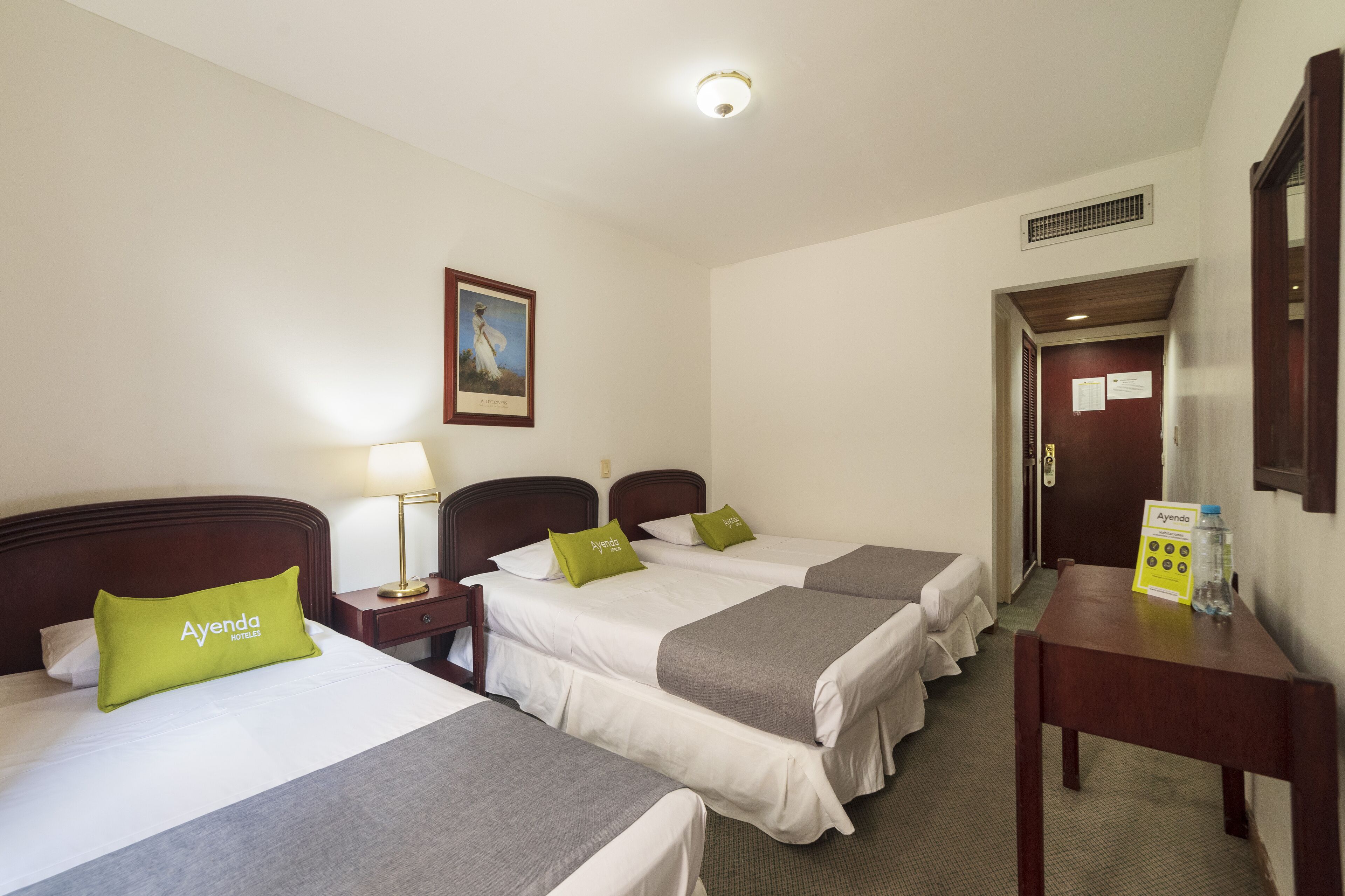 A hotel room with three single beds, each with a pillow and a bedside table. There is a nightstand with a lamp, a framed picture on the wall, and a door leading to another room.