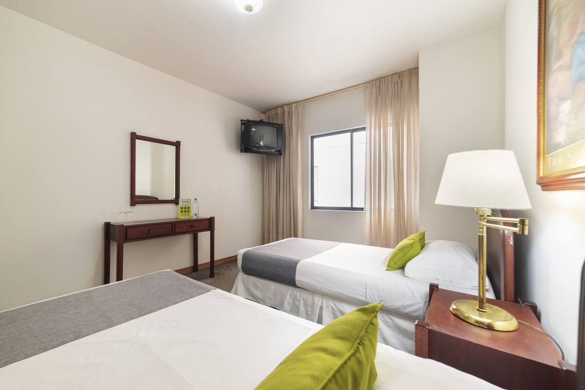 A hotel room with two beds, a bedside table with a lamp, a TV mounted on the wall, and a window with curtains.