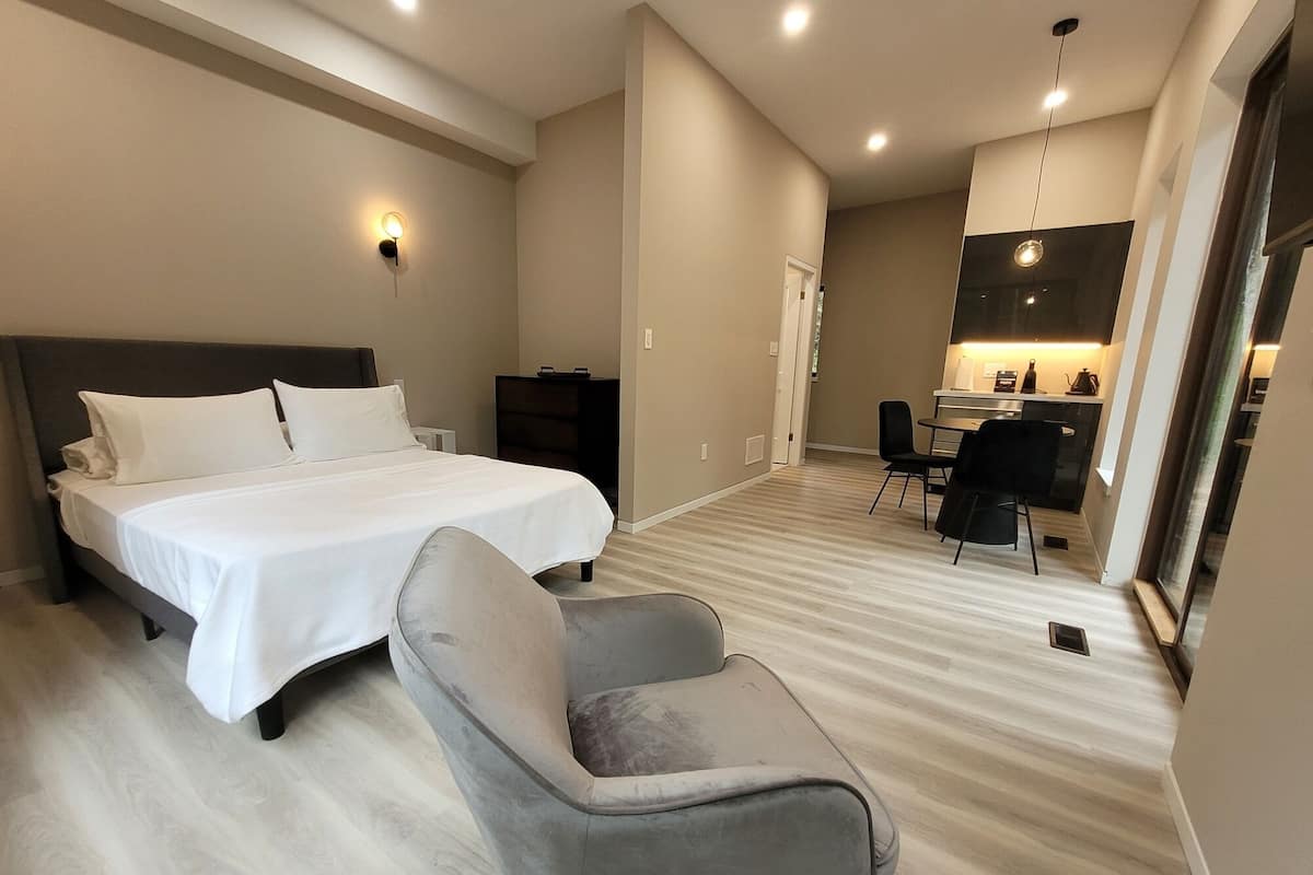 Quarto de hotel moderno com uma cama grande, uma poltrona cinza, uma escrivaninha com cadeira e uma cozinha compacta.