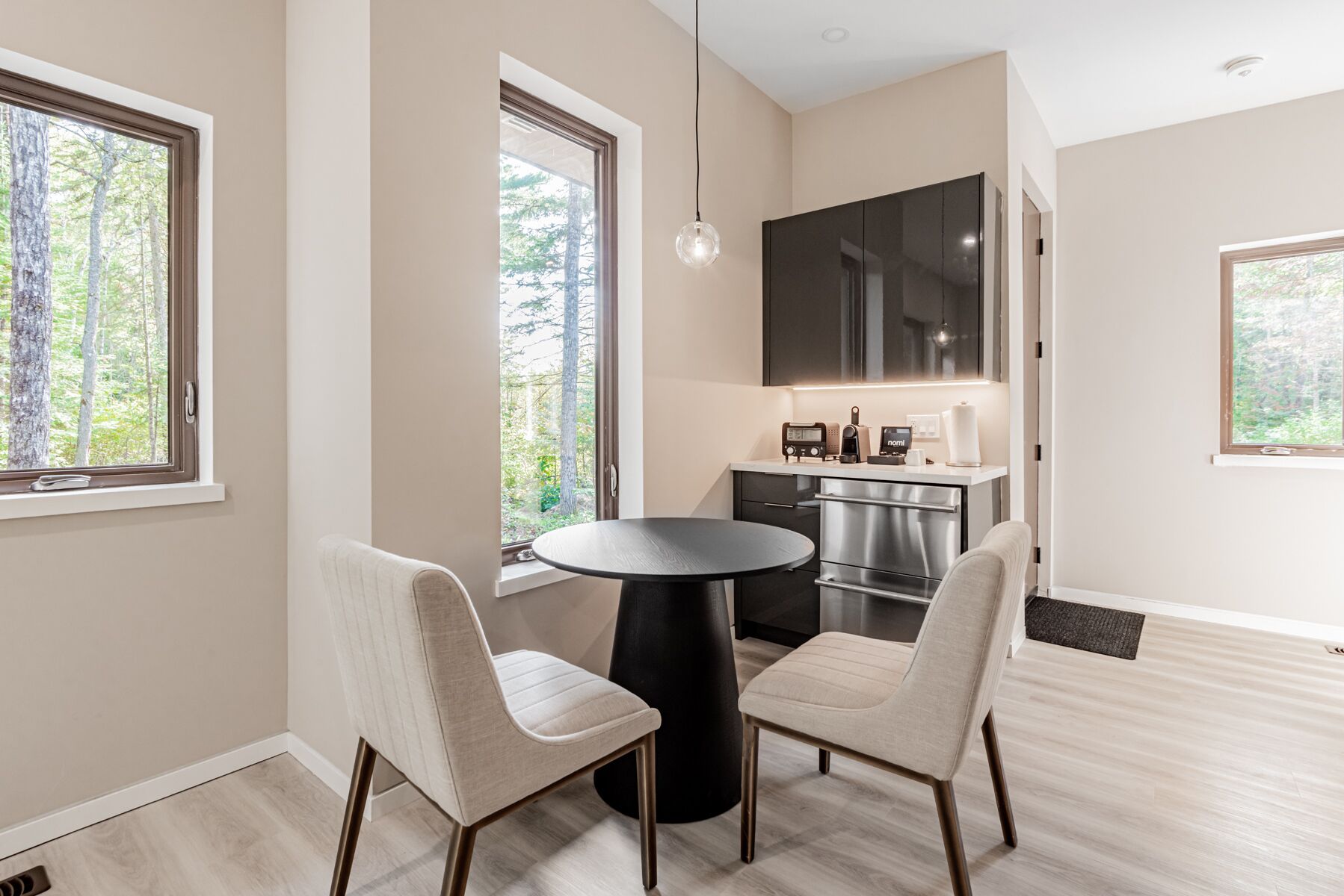A modern kitchen with a large TV, a dining area with two chairs and a round table, and large windows with a forest view.