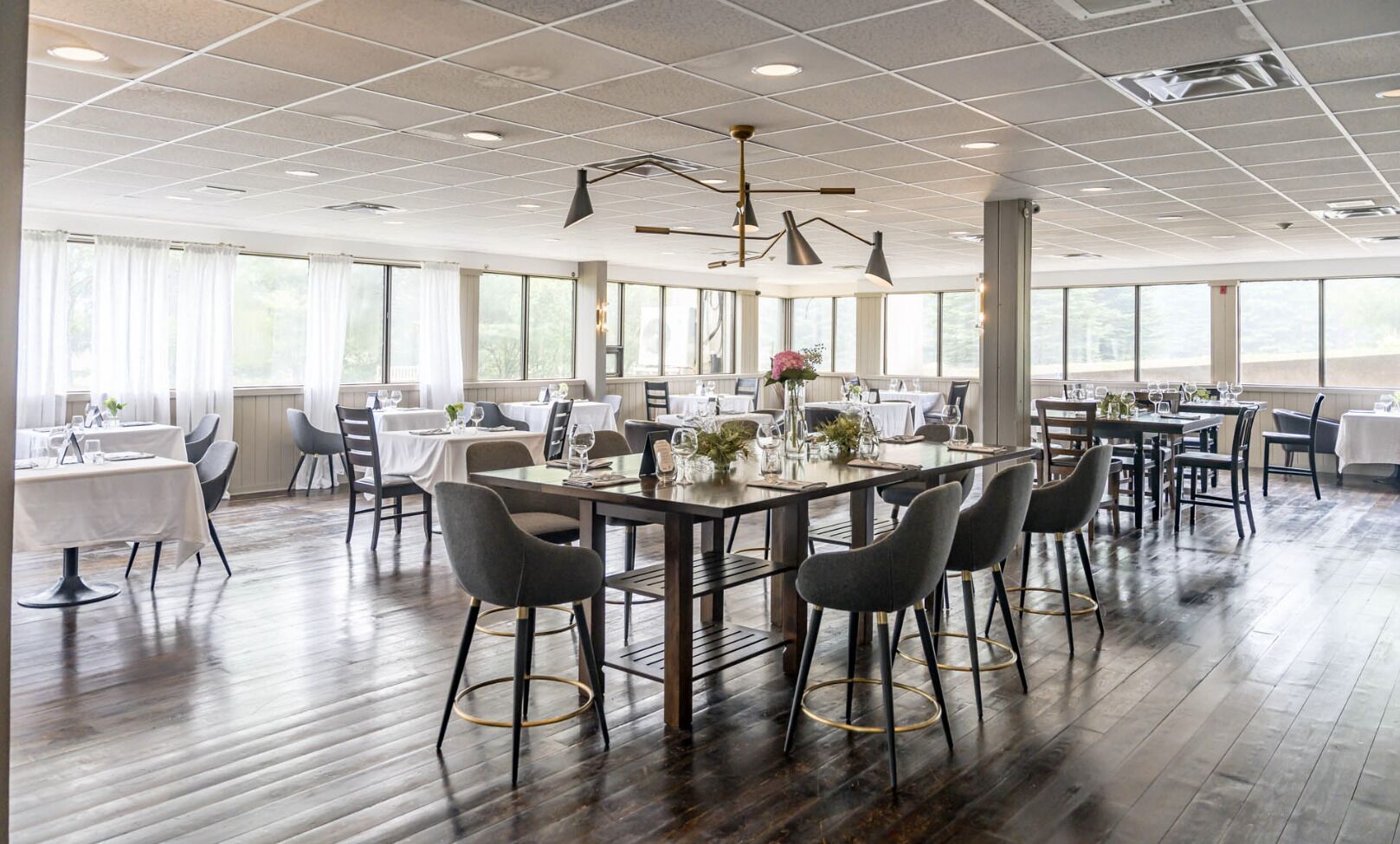 Uma sala de jantar ampla, com várias mesas e cadeiras, grandes janelas e um design moderno no teto.