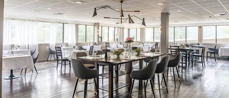Uma sala de jantar ampla, com várias mesas e cadeiras, grandes janelas e um design moderno no teto.