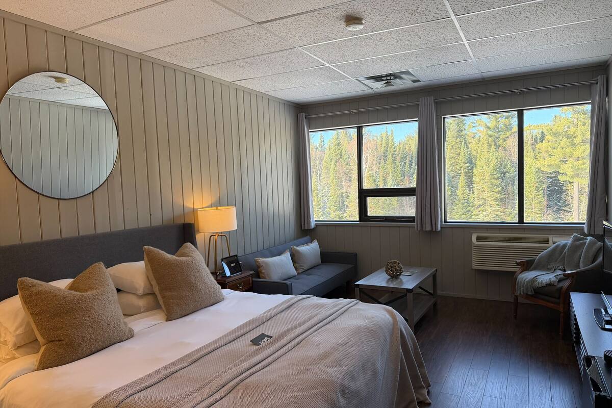 Quarto de hotel com uma cama grande, um sofá, uma mesa pequena, uma cadeira e uma janela vista para uma floresta.