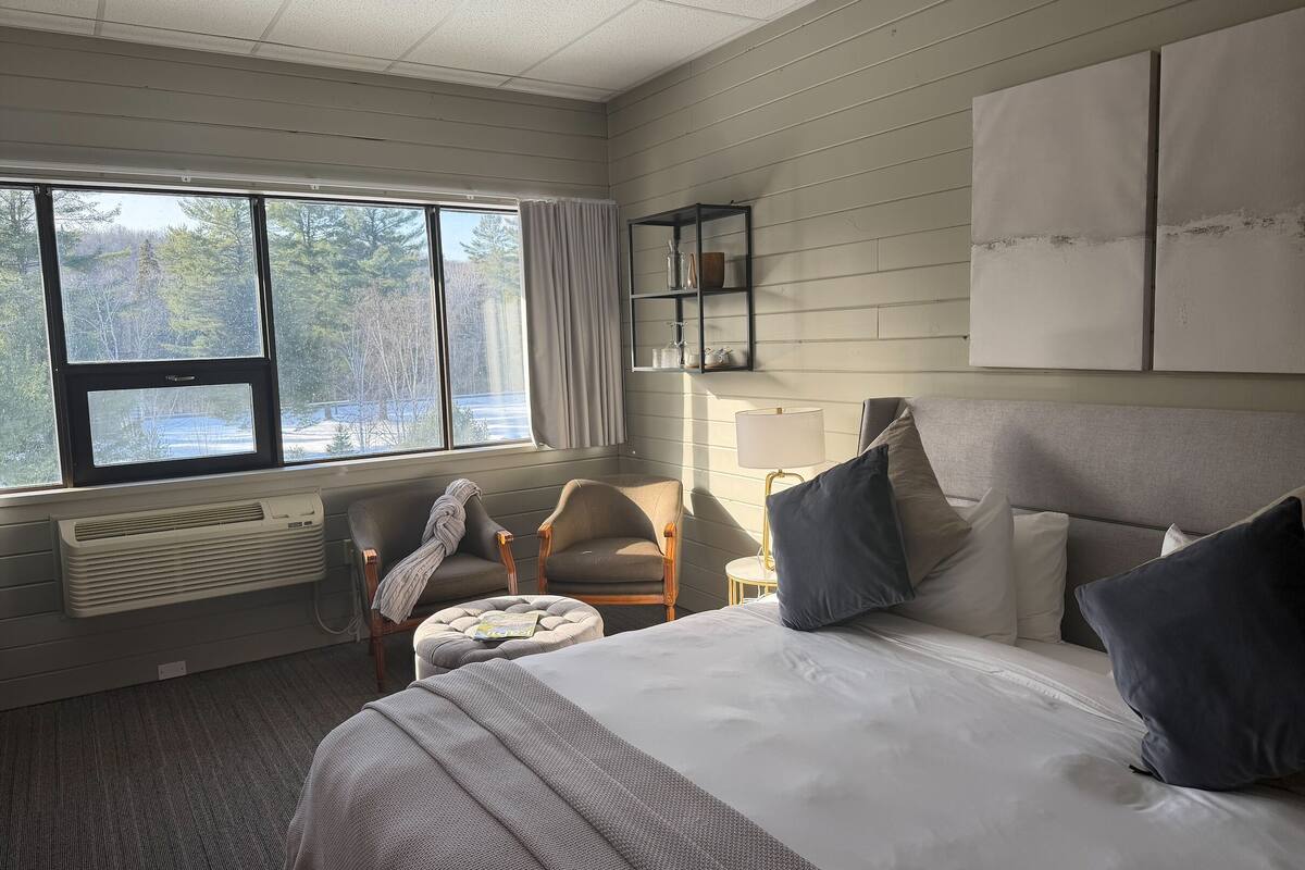 Quarto de hotel com uma cama grande, uma cadeira, uma mesinha e uma janela com vista para árvores.