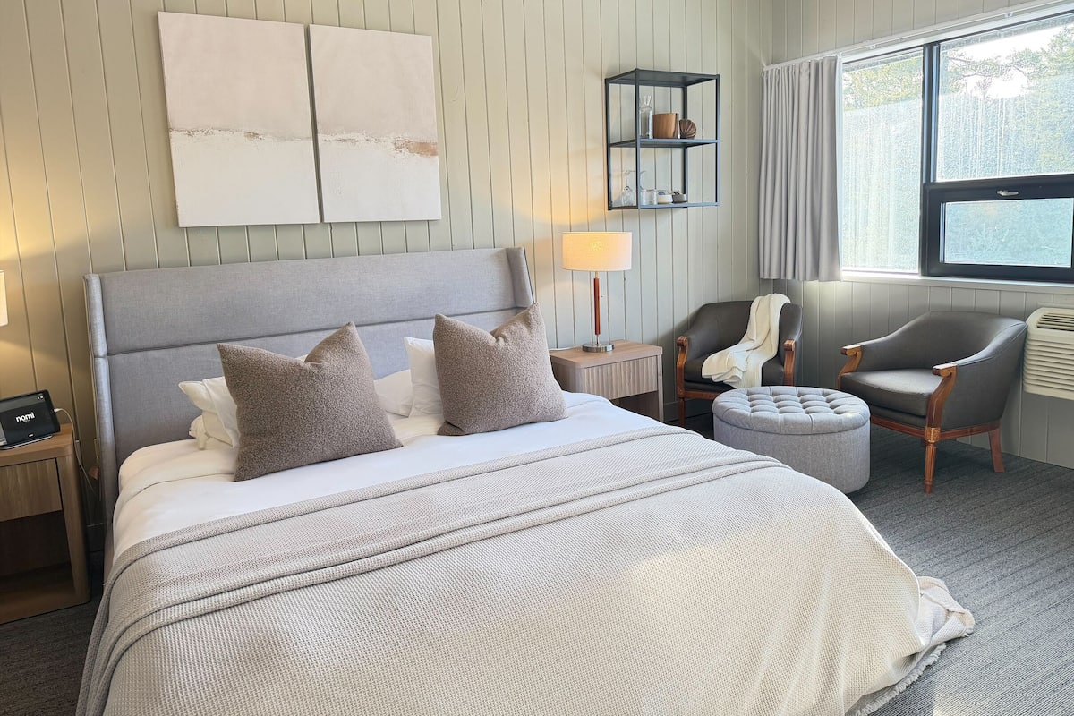 Quarto de hotel com uma cama grande, mesa de apoio, cadeira, um pequeno banquinho, mesa de cabeceira e janela com cortinas.