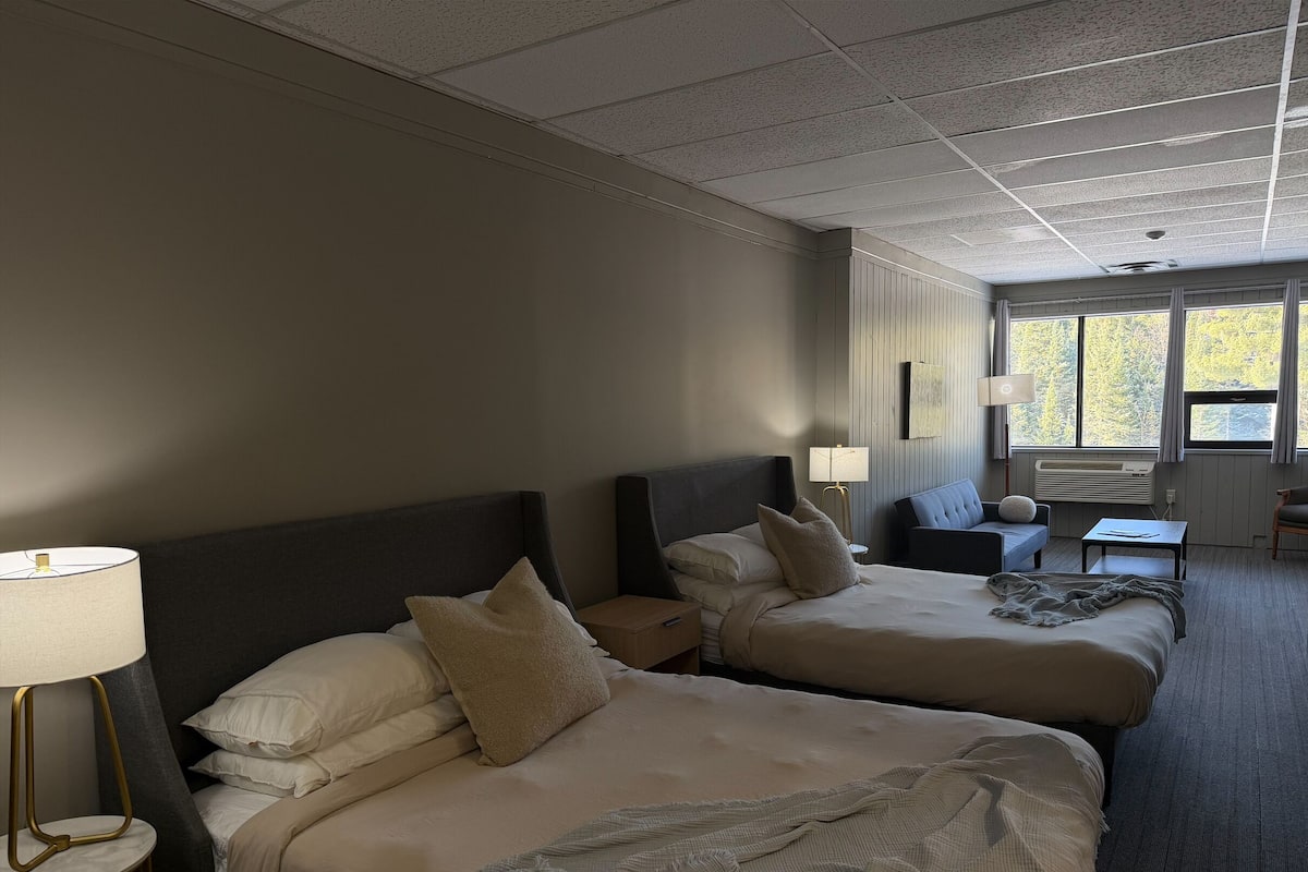 Quarto de hotel com duas camas, uma escrivaninha, uma cadeira, uma luminária e uma janela com vista para árvores.