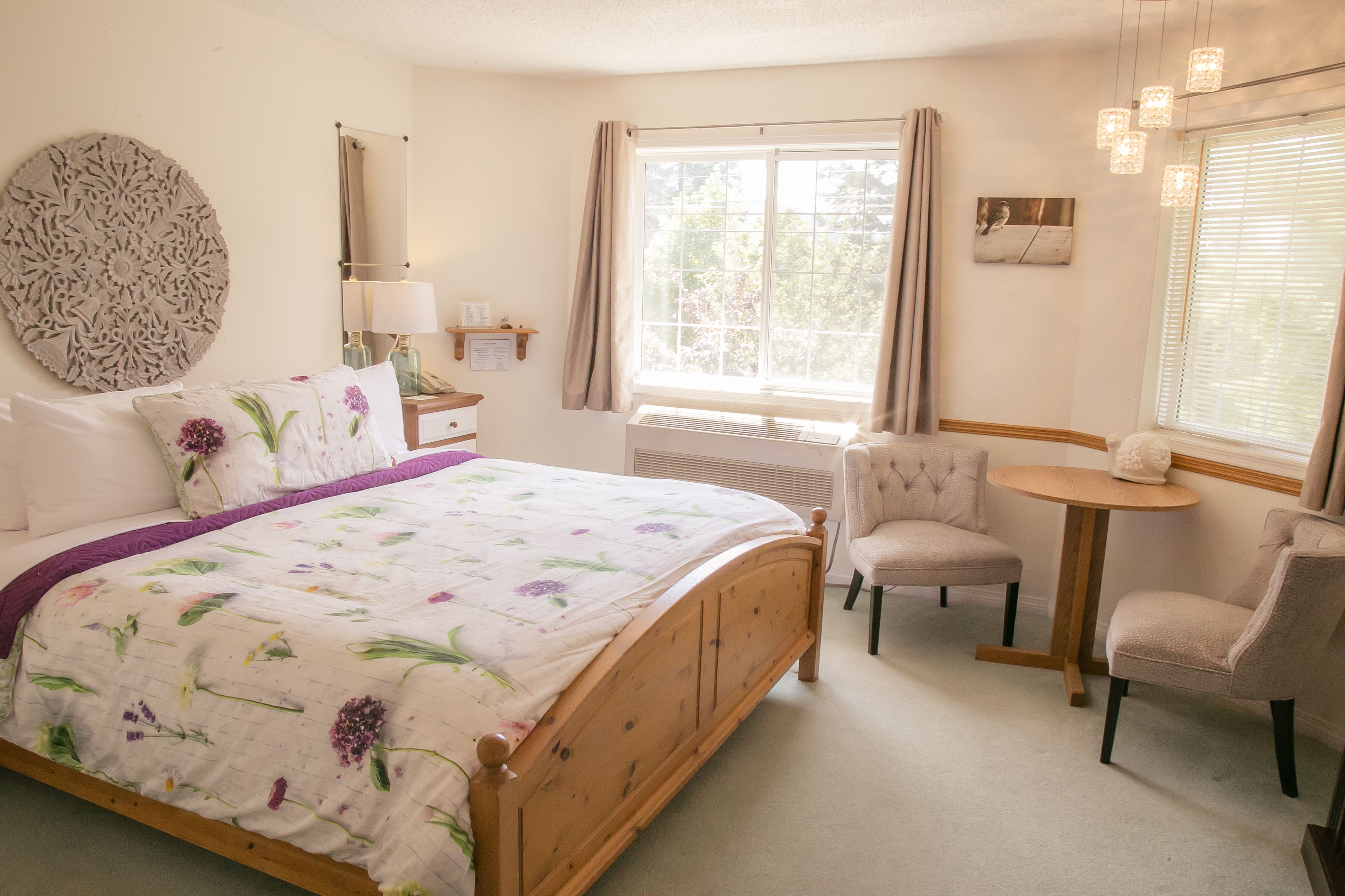 A bedroom with a wooden bed, a round table, two chairs, a window with curtains, a wall-mounted mirror, and a decorative wall piece.