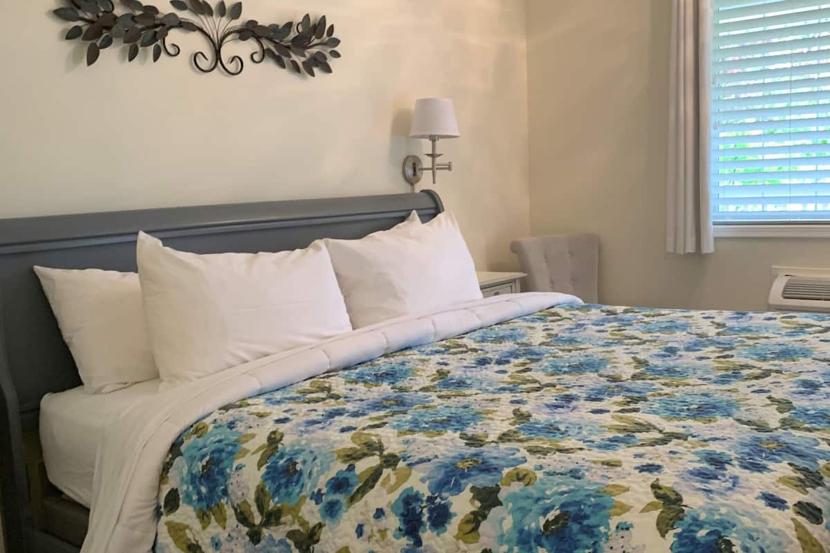 A neatly made bed with white linens and a blue floral bedspread, a wall-mounted decorative leaf art piece, a bedside lamp, and a window with blinds.