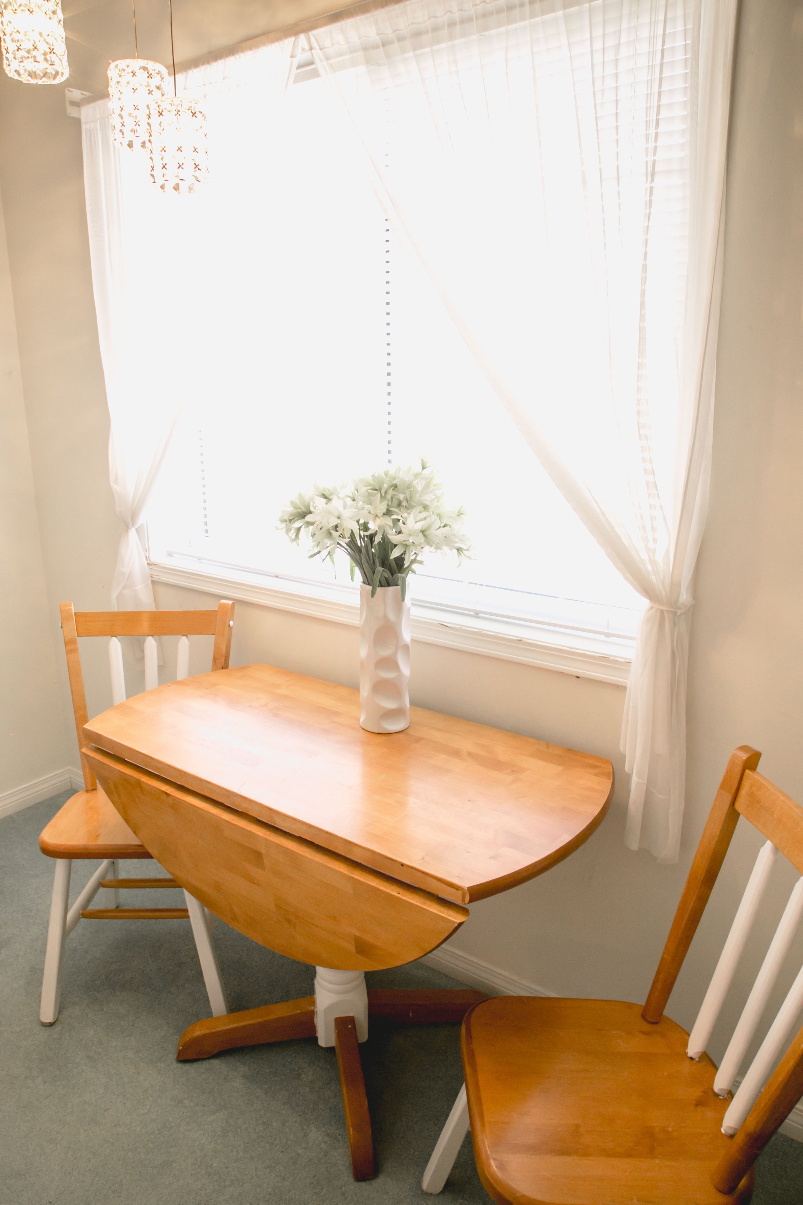 Una mesa de madera para comer con dos sillas, un florero con flores y una ventana con cortinas de gasa.