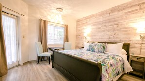 Camera da letto con testiera in legno, un letto con lenzuola a motivi floreali, una sedia, un comodino con una lampada e una finestra con tende.