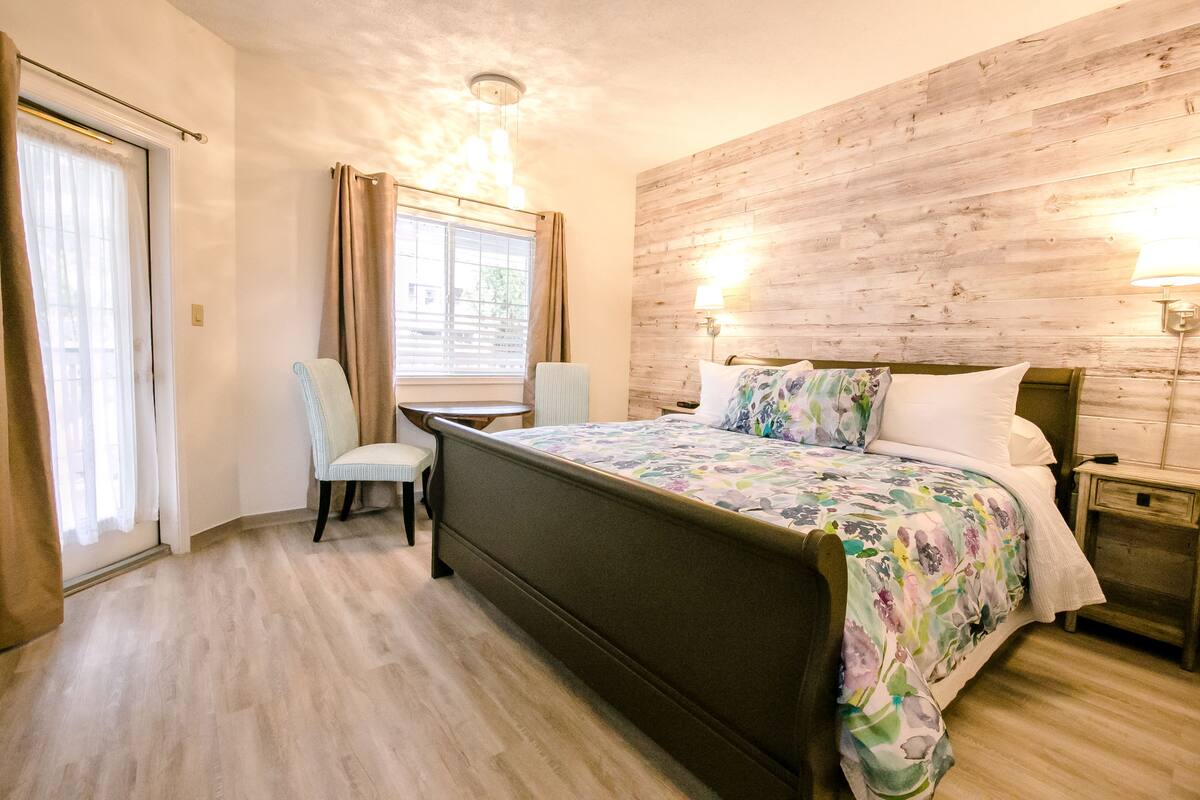 A bedroom with a wooden headboard, a bed with floral bedding, a chair, a nightstand with a lamp, and a window with curtains.