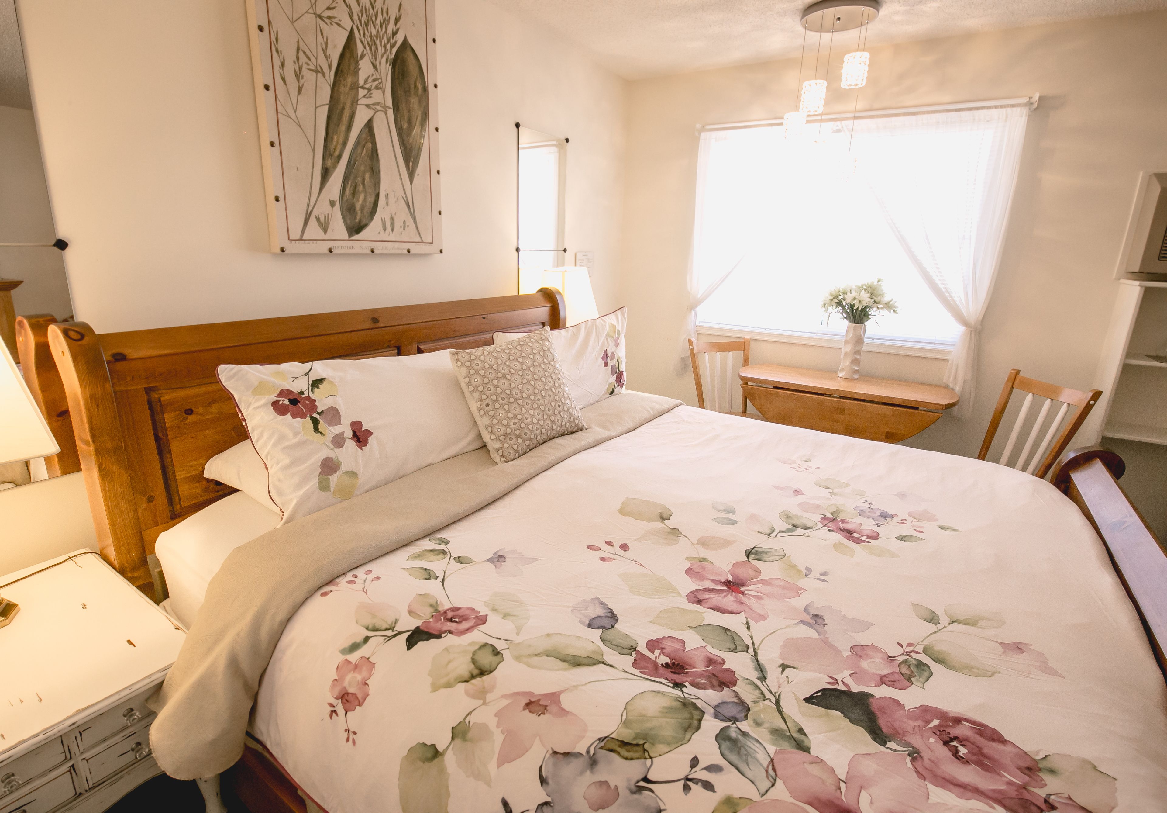 A bedroom with a wooden bed, floral bedding, a bedside table with a lamp, a window with sheer curtains, and a wall-mounted mirror.