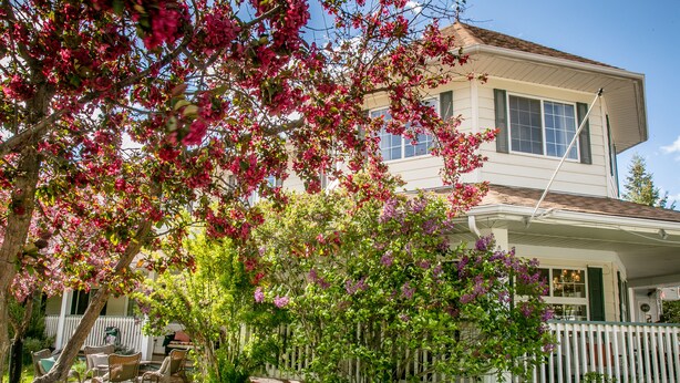 Una casa a due piani con facciata bianca, un portico con una staccionata bianca e un giardino fiorito.