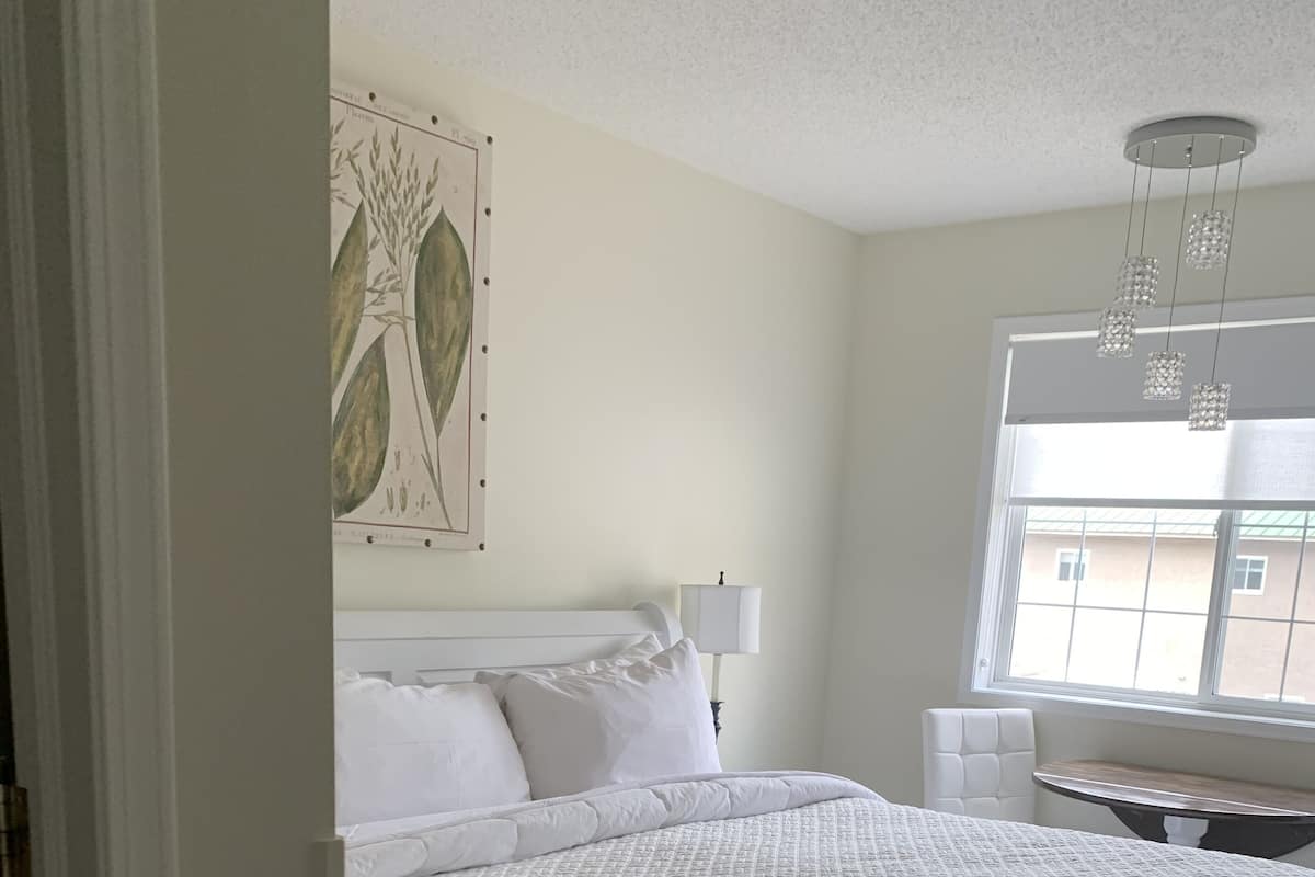 A neatly made bed with white linens in a room with a window, a small table, and a wall-mounted lamp.