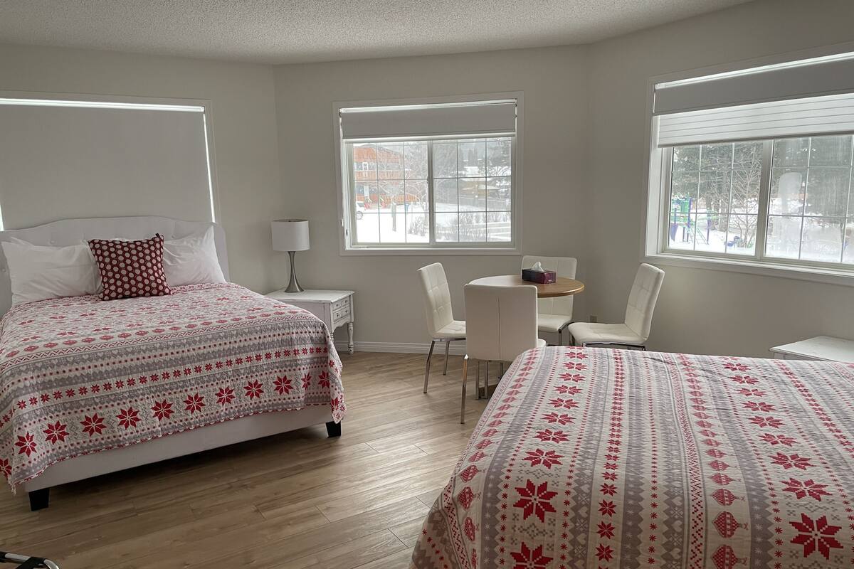 A hotel room with two beds, a small table, a chair, and a window.