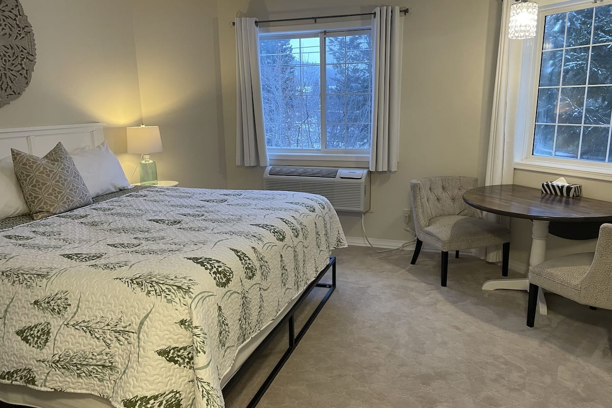 A bedroom with a bed, bedside table, lamp, air conditioning unit, window with curtains, and a small round table with chairs.