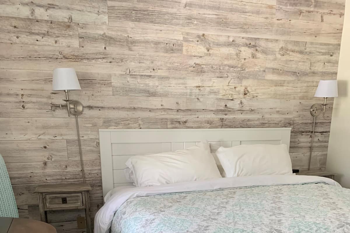 A bedroom with a wooden wall, a white bed, a bedside table, a chair, and two wall-mounted lamps.