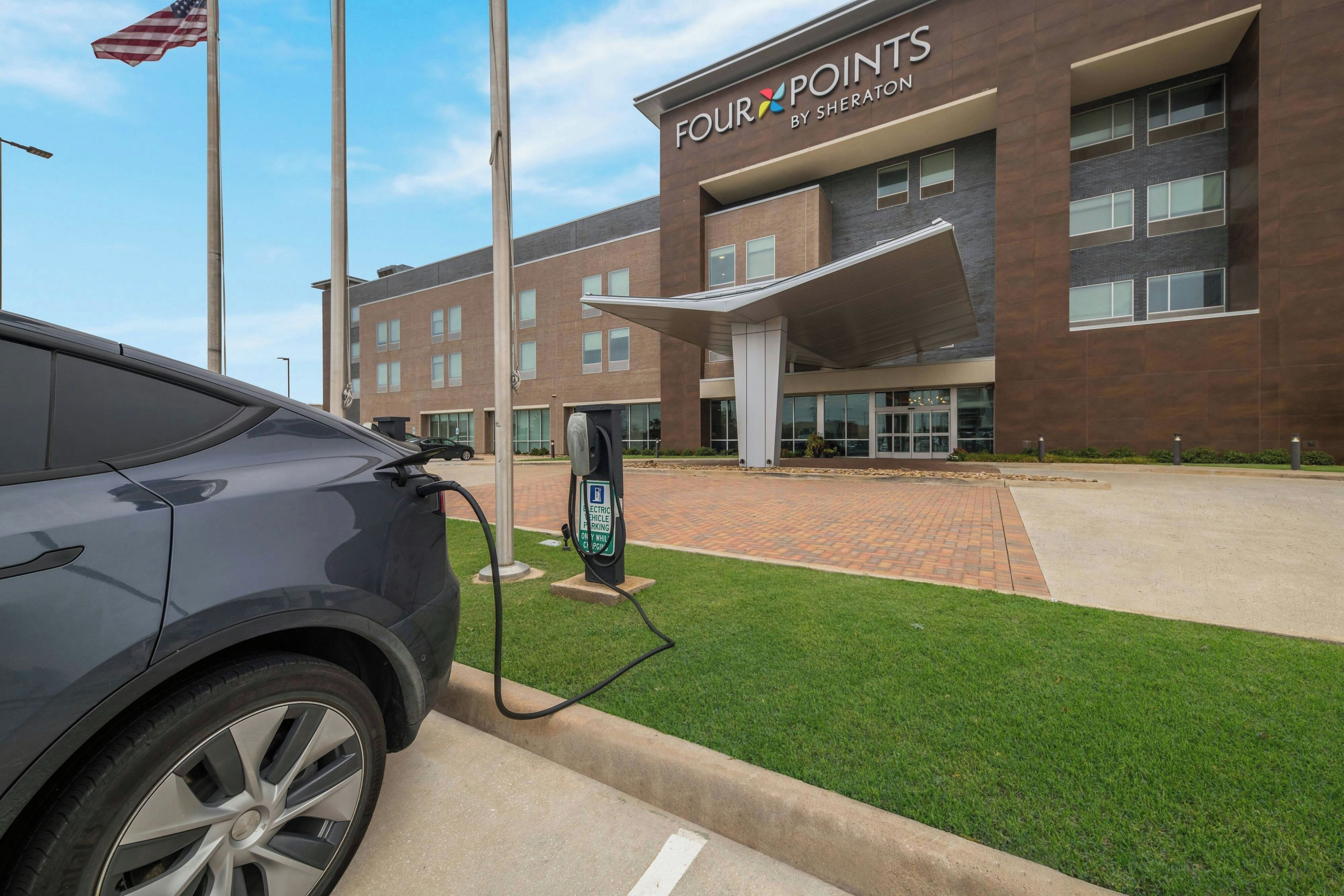 Un bâtiment moderne avec une enseigne indiquant « FOUR POINTS BY SHERATON » et une borne de recharge pour véhicules électriques devant l’entrée.